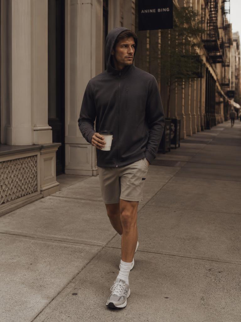 Model walking a quiet street in a charcoal performance hoodie with hood up and stone shorts